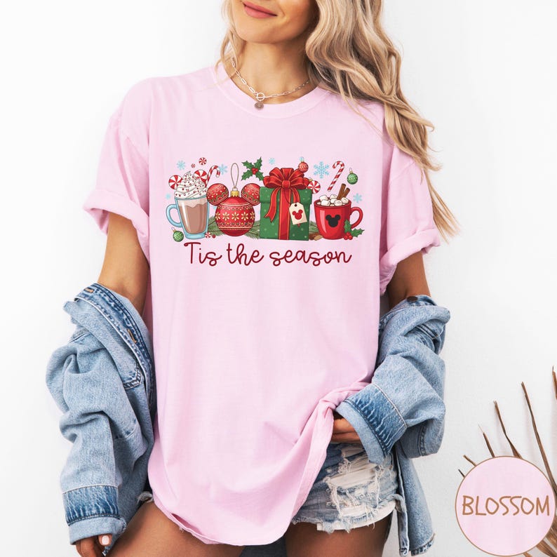 May include: A light pink t-shirt featuring a festive graphic with a hot chocolate, Christmas ornaments, a wrapped gift, and a mug. The text "Tis the season" is written in a script font. The design includes candy canes and snowflakes.