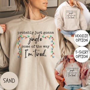 May include: Sand-colored sweatshirt with the text "probably just gonna jingle some of the way I'm tired" in black script, framed by a string of colorful Christmas lights. The image also shows a hoodie and t-shirt with the same design.