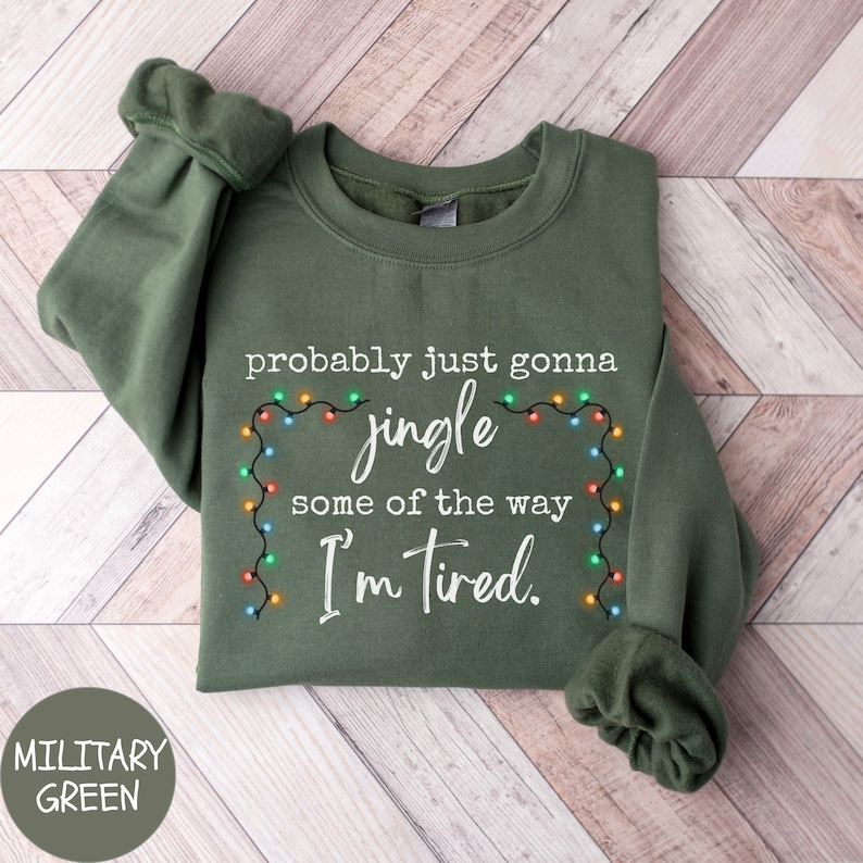 May include: Military green sweatshirt with the text "probably just gonna jingle some of the way I'm tired." The text is surrounded by a string of colorful Christmas lights. The sweatshirt is folded on a wooden surface.