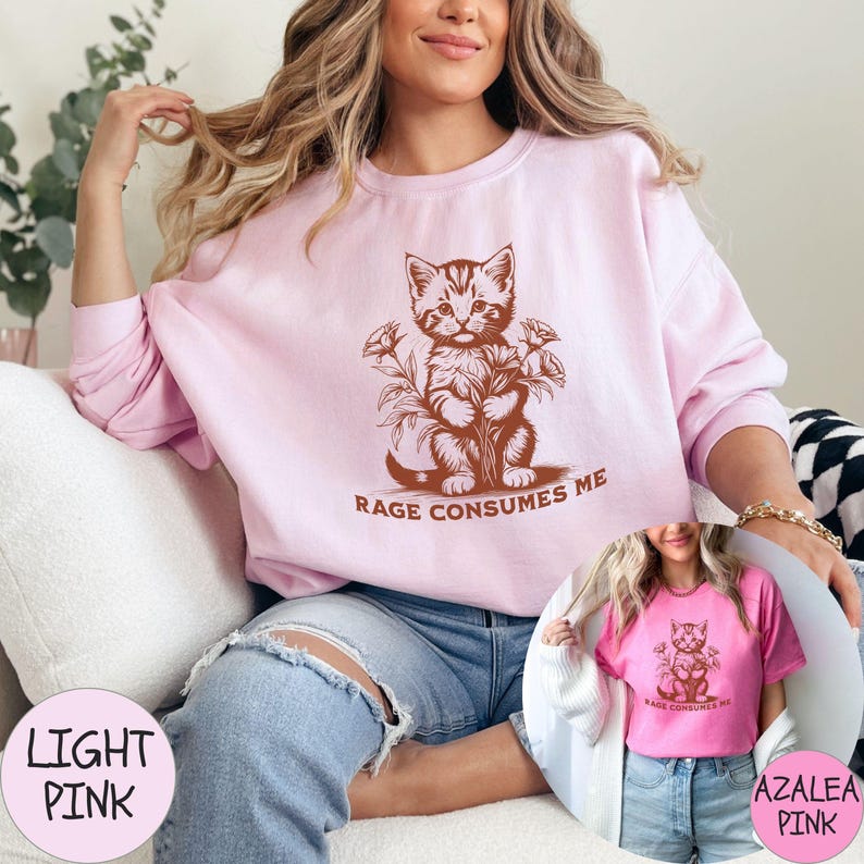 Puede incluir: Una sudadera rosa claro con una ilustraci&oacute;n marr&oacute;n de un gato sosteniendo flores y el texto "Rage Consumes Me".