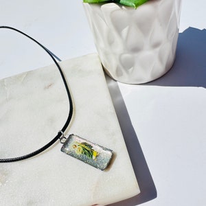 May include: A black cord necklace with a rectangular pendant featuring a colorful image of a person. The pendant is set in a clear resin with glitter.