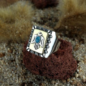 Op de afbeelding: Een zilveren ring met een rechthoekige voorkant met een turquoise ovale steen en een gestileerd vogelontwerp. De ring heeft een crèmekleurige achtergrond en een zwarte rand. De ring ligt op een rode steen.