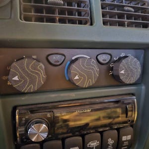 May include: Close-up of a vehicle's dashboard featuring a Pioneer stereo and climate control knobs. The knobs have a gray and yellow topographic design. The dashboard is olive green.