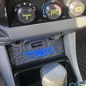 May include: Interior car detail featuring a center console with a custom insert. The insert has a gray topographic pattern and the blue letters "TRD". The dashboard has climate control knobs and a carbon fiber trim.