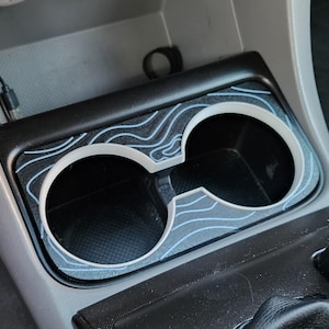 May include: A black car cup holder with a white and blue topographic design liner. The cup holder is empty.