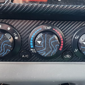 Könnte beinhalten: Nahaufnahme eines Armaturenbretts eines Autos mit fünf runden Knöpfen. Die Knöpfe sind schwarz mit einem blau-weißen Wellenmuster. Die Knöpfe sind mit "OFF", "LO", "HI", "TEMP" und "MAX A/C" beschriftet.