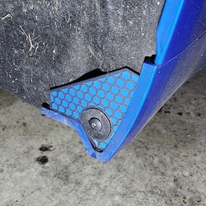 Puede incluir: Primer plano de un componente azul de coche con un patrón de panal de abeja negro y azul. El componente está fijado con un sujetador negro. El fondo es una superficie oscura y texturizada.