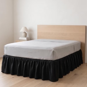 May include: A black bed skirt with a gathered ruffle design, positioned around the base of a bed frame. The bed features a light gray fitted sheet and is set against a light wood headboard in a room with a lamp on a nightstand.