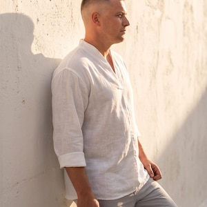 May include: A long-sleeved white shirt with a V-neckline and rolled cuffs, paired with light gray trousers. The shirt features a textured fabric and a relaxed fit, worn untucked. The overall appearance is clean, simple, and neutral in its design.