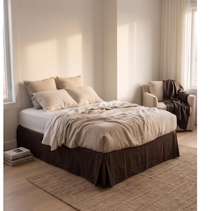 May include: A bed featuring a white fitted sheet, a light beige duvet cover, and four matching beige pillows. The bed base is covered by a dark brown bed skirt. An armchair with a dark brown throw blanket sits nearby on a woven rug.