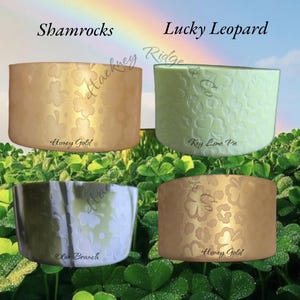 Puede incluir: Cuatro recipientes decorativos con diseños de tréboles y leopardo. Los recipientes son en tonos dorados, verdes y blancos. El texto en la imagen dice "Shamrocks" y "Lucky Leopard". Los recipientes están sobre un fondo de hojas de trébol y un arcoíris.