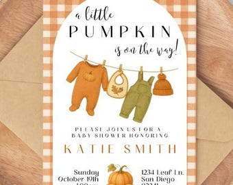May include: Baby shower invitation with a pumpkin theme. The invitation features the text "a little PUMPKIN is on the way!" and illustrations of baby clothes. The invitation is on a wooden tray with a brown envelope.