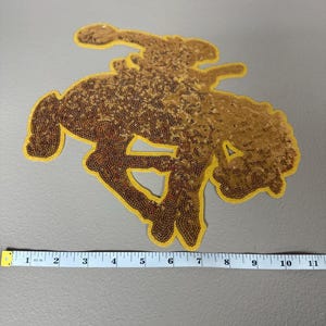 May include: A gold sequined patch in the shape of a rodeo rider on a bucking bronco. The patch is outlined in yellow and measures approximately 10 inches in length.
