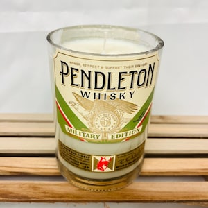 May include: A glass candle jar with a white candle inside. The jar is decorated with a label that says "Pendleton Whisky Military Edition". The label is gold, green, and red with an eagle and a star.