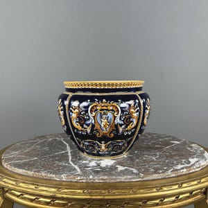 May include: A dark blue ceramic planter featuring intricate gold, white, and light blue scrollwork and cherub motifs. The rim is decorated with a repeating yellow and white pattern. The vessel has a rounded, bulbous shape with a glossy finish.