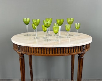 May include: An oval marble top table with a wood base and carved details. The table is decorated with a set of green glass goblets.