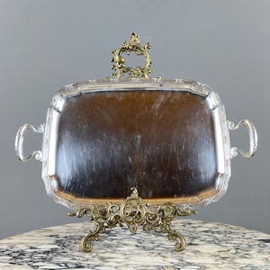 Puede incluir: Una bandeja rectangular plateada con asas ornamentadas y una tapa decorativa. La bandeja está sostenida por un soporte dorado de diseño intrincado. La bandeja se exhibe sobre una superficie con vetas de mármol.