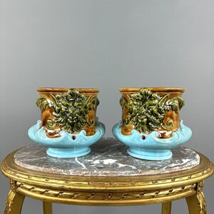 Peut inclure: Une paire de cache-pots en céramique présentant un motif de visage vert en relief sur un corps brun, posés sur une base bleu clair avec un petit accent rouge. Chaque cache-pot a un bord évasé et une finition brillante, avec des détails sculptés.