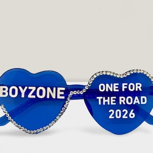 Op de afbeelding: Blauwe hartvormige zonnebril met de tekst "BOYZONE" op de linkerlens en "ONE FOR THE ROAD 2026" op de rechterlens. De randen zijn versierd met kleine, heldere strass-steentjes.
