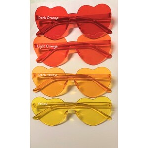 May include: Four pairs of heart-shaped sunglasses in shades of orange and yellow. The top pair is dark orange, followed by light orange, dark yellow, and yellow. Each pair has a transparent frame and lenses, with the color names printed above.