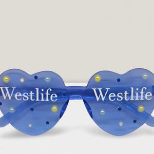 May include: Blue heart-shaped sunglasses with the word "Westlife" in white lettering. The sunglasses have decorative yellow, blue, and clear embellishments. The frame is a transparent blue colour.