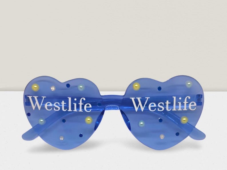 May include: Blue heart-shaped sunglasses with the word "Westlife" in white lettering. The sunglasses have decorative yellow, blue, and clear embellishments. The frame is a transparent blue colour.