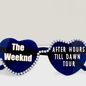 May include: Heart-shaped sunglasses in a dark blue and black plaid design. The text "The Weeknd" and "AFTER HOURS TILL DAWN TOUR" are in white. The frames are edged with small white beads.