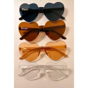 May include: Four pairs of heart-shaped sunglasses in black, brown, tan, and clear. The sunglasses have a rimless design and are arranged vertically. The text "Black", "Brown", "Tan", and "Clear" are written on the image.