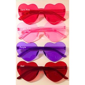 May include: Four pairs of heart-shaped sunglasses in shades of dark pink, light pink, purple, and red. Each pair has a rimless design with a heart-shaped lens and matching arms. The text "Dark Pink", "Light Pink", "Purple", and "Red" are visible.