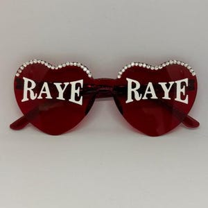 May include: Red heart-shaped sunglasses with the word "RAYE" in white lettering on each lens. The rims are adorned with small, sparkling rhinestones. The sunglasses have a classic, stylish design.