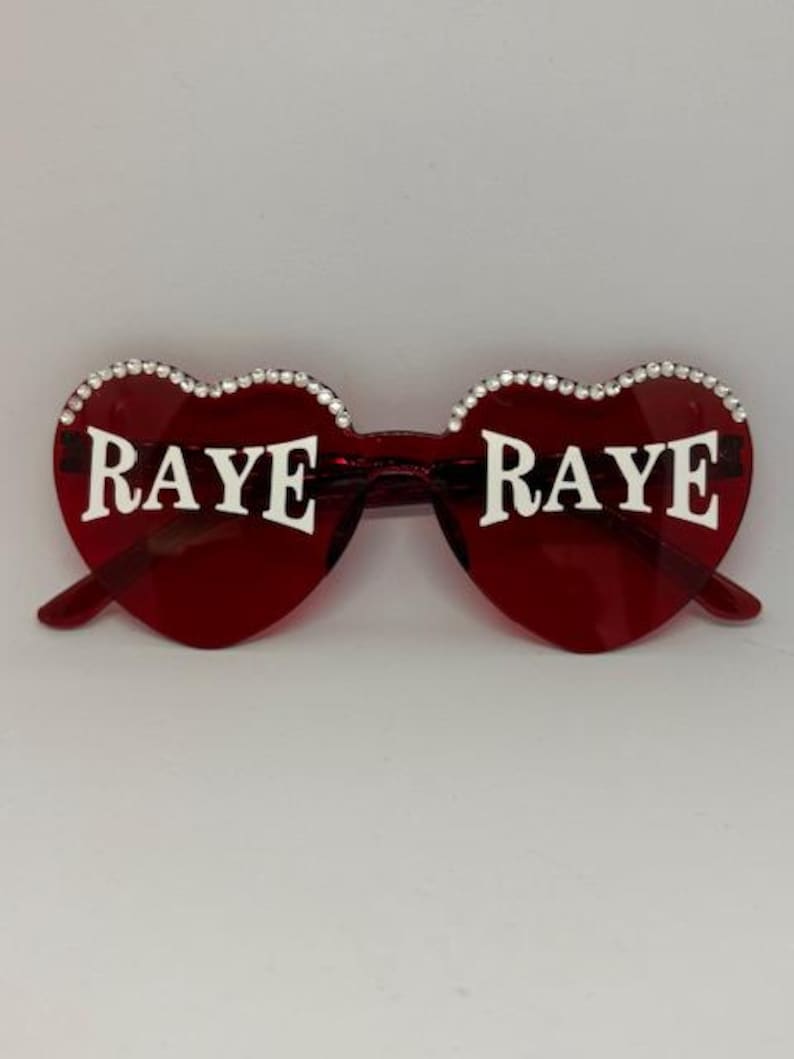 May include: Red heart-shaped sunglasses with the word "RAYE" in white lettering on each lens. The rims are adorned with small, sparkling rhinestones. The sunglasses have a classic, stylish design.