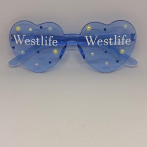 May include: Blue heart-shaped sunglasses with the word "Westlife" printed in white on each lens. The sunglasses are decorated with small, round, pearl-like embellishments. The frame is a transparent blue colour.