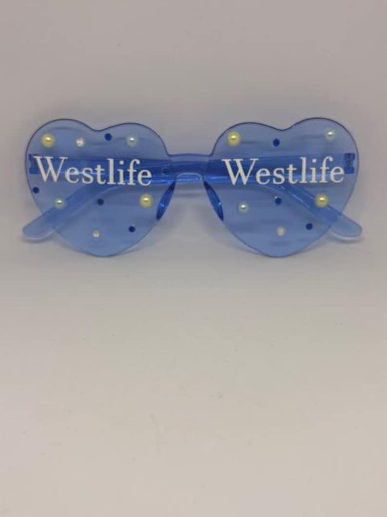 May include: Blue heart-shaped sunglasses with the word "Westlife" printed in white on each lens. The sunglasses are decorated with small, round, pearl-like embellishments. The frame is a transparent blue colour.