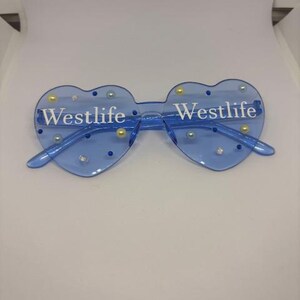 May include: Blue heart-shaped sunglasses with the word "Westlife" printed in white on each lens. The sunglasses are decorated with small, round, pearl-like embellishments. The frame and arms are a matching blue colour.