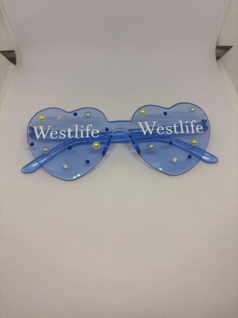 May include: Blue heart-shaped sunglasses with the word "Westlife" printed in white on each lens. The sunglasses are decorated with small, round, pearl-like embellishments. The frame and arms are a matching blue colour.