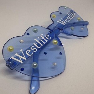 May include: Blue heart-shaped sunglasses with the word "Westlife" printed in white. The sunglasses have a clear frame and are decorated with pearls and rhinestones. The lenses are a light blue colour.