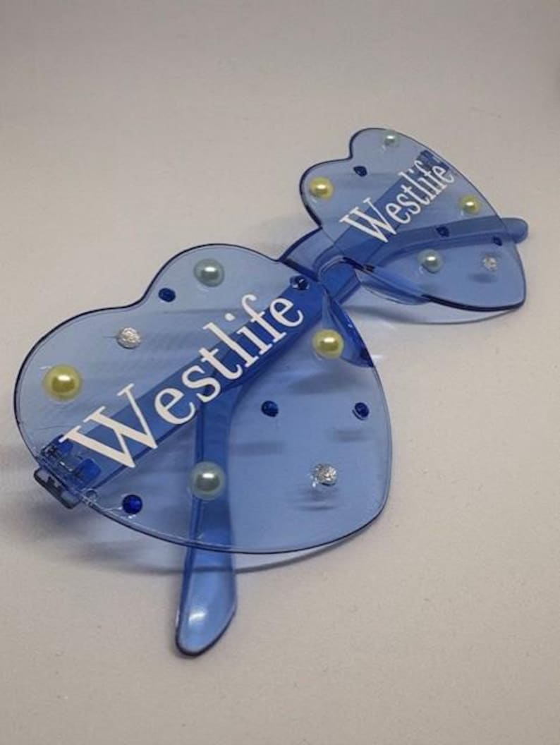 May include: Blue heart-shaped sunglasses with the word "Westlife" printed in white. The sunglasses have a clear frame and are decorated with pearls and rhinestones. The lenses are a light blue colour.