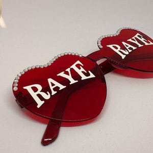 May include: Red heart-shaped sunglasses with the word "RAYE" in white lettering. The frames are a vibrant red, and the edges are adorned with small, sparkling embellishments. The sunglasses have a modern, stylish design.