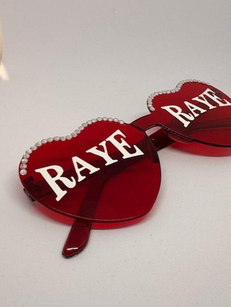 May include: Red heart-shaped sunglasses with the word "RAYE" in white lettering. The frames are a vibrant red, and the edges are adorned with small, sparkling embellishments. The sunglasses have a modern, stylish design.