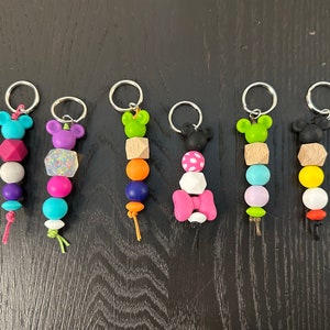 Op de afbeelding: Zeven kleurrijke sleutelhangers met verschillende figuurtjes en kralen. De figuurtjes zijn allemaal cartoonfiguren met een Mickey Mouse-thema. De kralen zijn in verschillende kleuren, waaronder roze, paars, groen, blauw, geel en oranje.