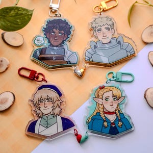 Può includere: Quattro portachiavi con personaggi in acrilico con chiusure in metallo. I personaggi sono raffigurati in armatura e abbigliamento fantasy. I portachiavi presentano un personaggio dai capelli scuri con una nuvoletta, un personaggio biondo in armatura, un personaggio con un cappello blu e un personaggio con trecce.