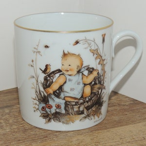 Può includere: Tazza in porcellana bianca con bordo dorato. La tazza presenta un'illustrazione colorata di un bambino seduto in un cesto con un uccello e fiori.