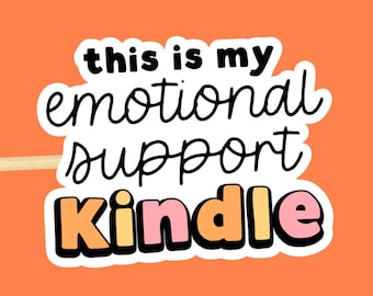 Puede incluir: Una pegatina troquelada con el texto negro "this is my emotional support" sobre la palabra "Kindle" en letras grandes y coloridas con tonos naranja, amarillo y rosa, todo delineado en blanco sobre un fondo naranja sólido.