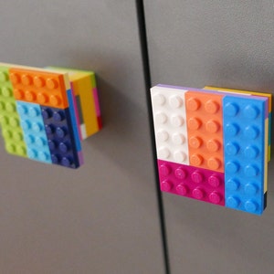 May include: Two colourful Lego brick magnets attached to a grey surface. One magnet features green, orange, blue, and purple bricks. The other magnet has white, orange, pink, and blue bricks. These magnets are a fun and playful addition to any space.