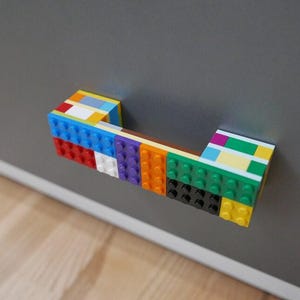Può includere: Una maniglia colorata per armadi realizzata con mattoncini giocattolo ad incastro. La maniglia presenta una varietà di colori, tra cui blu, rosso, giallo, verde e viola, attaccata a una porta di un armadio grigio. La maniglia è lunga circa 15 cm.
