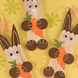 May include: Handmade Easter bunny crafts made from popsicle sticks, featuring brown ears, googly eyes, and orange carrot accents. The bunnies have brown circular feet and are set against a yellow background with floral cutouts.