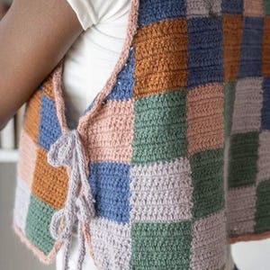 Könnte beinhalten: Eine bunte gehäkelte Weste mit Patchwork-Design. Die Weste hat Quadrate in verschiedenen Farben, darunter Blau, Orange, Rosa, Grün und Grau. Die Kanten sind mit einem rosa Rand versehen und haben Bindebänder.