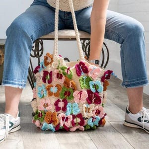 Könnte beinhalten: Eine cremefarbene Häkeltasche mit bunten Schmetterlingsapplikationen in Rosa, Blau, Grün und Orange. Die Tasche hat zwei cremefarbene Henkel. Die Person trägt eine blaue Jeans und weiße Turnschuhe.