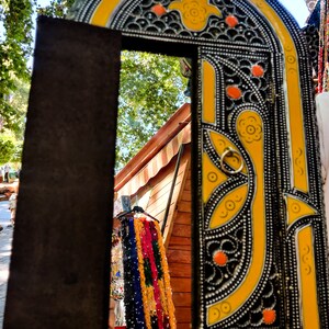 May include: A decorative mirror with a yellow and black frame. The frame is intricately carved with floral patterns and has a small, round, yellow hand-shaped design in the center. The mirror is reflecting a wooden structure with colorful fabric hanging from it.
