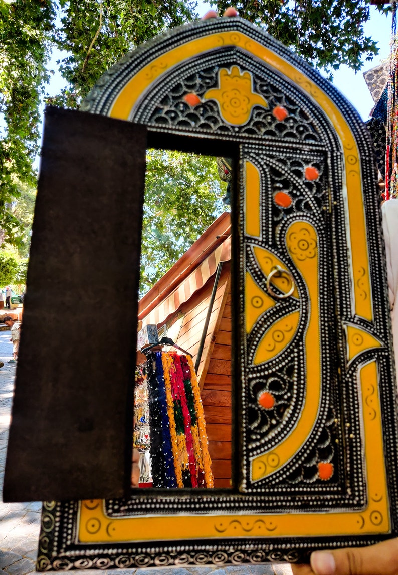 May include: A decorative mirror with a yellow and black frame. The frame is intricately carved with floral patterns and has a small, round, yellow hand-shaped design in the center. The mirror is reflecting a wooden structure with colorful fabric hanging from it.